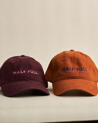 Two baseball caps, one maroon and one orange, with 'HALF FULL' text on a beige background.